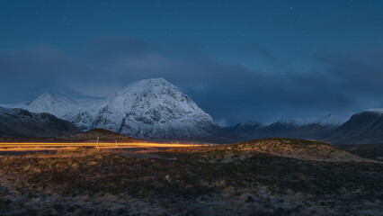 Night view of a road leading through the Scottish Highlands of Glen Coe with car light trail, snowcapped mountains and valley. Scotland - obrazy, fototapety, plakaty