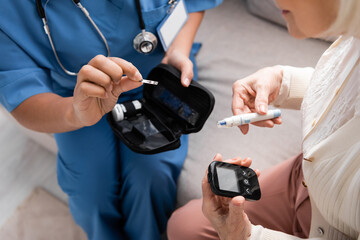 cropped view of multiracial nurse in uniform holding test strip near senior woman holding lancet pen and glucometer.