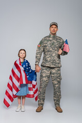 serviceman in military uniform holding flag of USA and hand of happy daughter on grey.