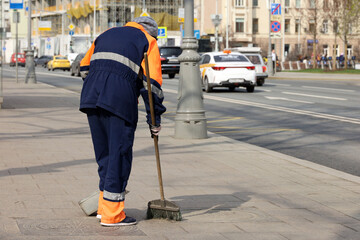 Woman worker in orange uniform with a broom sweeps the sidewalk on cars background. Street cleaning in spring city