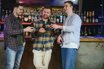 Three young men in casual clothes are smiling, holding bottles of beer while standing near bar counter in pub