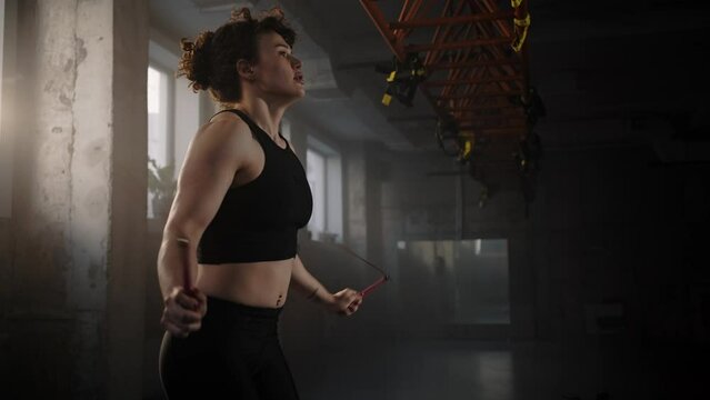 Side view of redhead Caucasian woman athlete weightlifter exercising with jumping rope in smokey gym