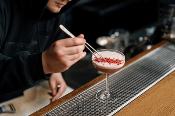 The bartender prepares a milk punch cocktail and puts chopped raspberries on top of the drink with tongs