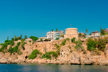 Fototapeta na wymiar View of the rocky coast with a tower and modern buildings on it from the sea in the city of Antalya.