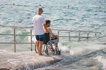 Man with disability at beach goes to swimm on a wheelchair with assistance on an accessible ramp.