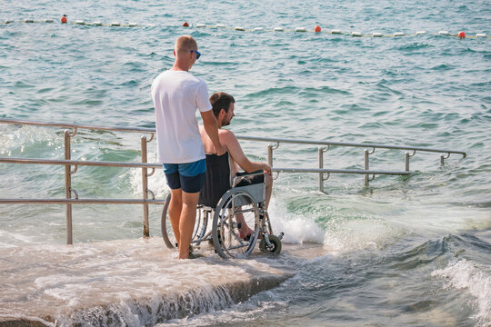 Man with disability at beach goes to swimm on a wheelchair with assistance on an accessible ramp.