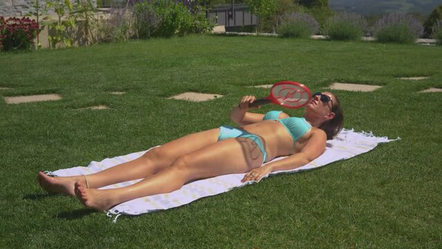 CLOSE UP: Pretty lady tries to enjoy sunbathing when insects start bothering her. Funny shot of a young woman flapping around with electric fly swatter to protect herself from pesky mosquitos.