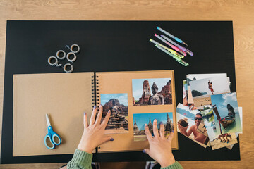 Image seen from above of a middle-aged woman's hands placing the photos and markers to make her handmade travel kraft paper photo album.