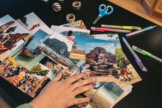 Hand of unrecognizable woman choosing which photo to pick from the pile of travel photos next to a brown kraft album ready to be made with whasi tape.