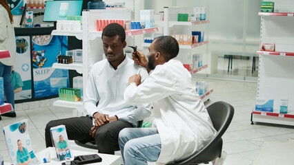 Health specialist consulting client with otoscope at clinic, sitting in pharmacy and doing ear examination. Young otologist and customer checking infection for otolaryngology consultation.