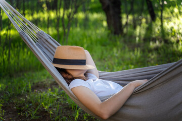 Woman resting in a hammock in a summer garden covering her face with a straw hat. Summer relax vacation - 594442389