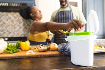 Happy senior african american couple wearing aprons and throwing waste out in kitchen