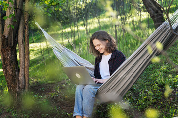 Young cheerful smiling woman working online using laptop sitting in hammock in summer garden