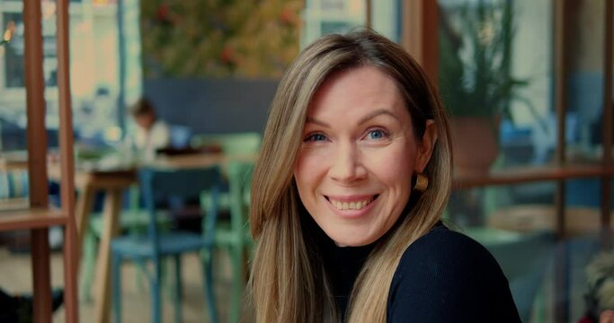 Head shot woman raise eyebrows smile openly in urban cafe with genuine interior. Straight camera look of cheerful lady with long light brown hair in city restaurant.