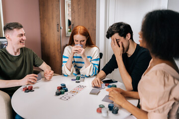 Happy excited multiethnic friends having home fun party on weekends playing poker game sitting at table in living room. Diverse men and women satisfied with spending leisure together playing poker.
