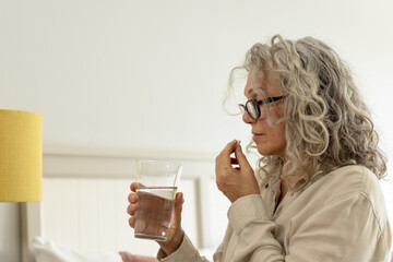 Senior caucasian woman wearing glasses sitting on bed, holding glass of water and pill