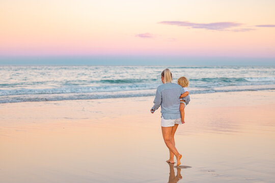 Single Mother Carrying Son On The Beach In Beautiful Twilight Light. Sunset On The Gold Coast, Queensland Australia