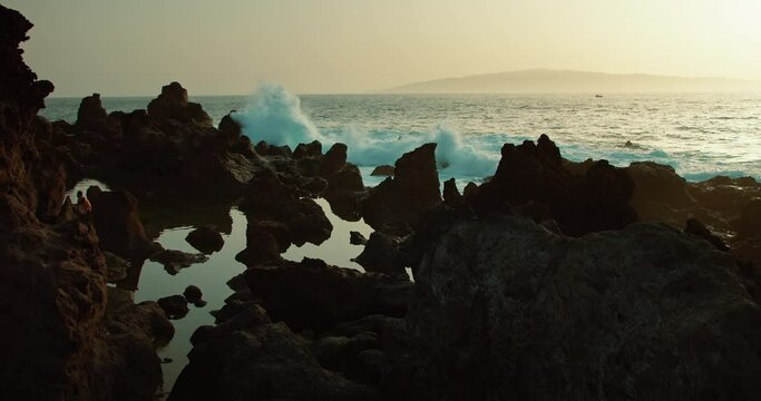 Powerful ocean waves breaking on dark sharp volcanic rocks at dusk. Devil's natural pool in Tenerife. Breathtaking view of sea water on rocky tropical shore.