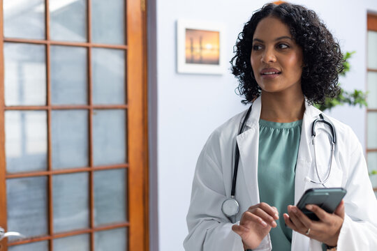 Biracial female doctor wearing sthethoscope, using smartphone at doctor's office