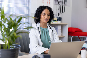 Biracial female doctor sitting at desk and using laptop at doctor's office