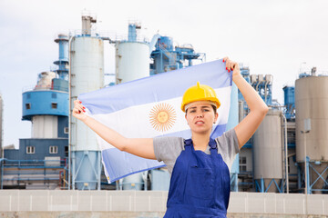 Sad unhappy young female worker in helmet waving national flag of Argentina during strike in front of big tanks at refinery factory