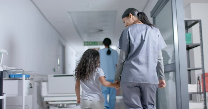 Diverse Female Nurse And Child Patient Walking Through Corridor At Hospital, In Slow Motion