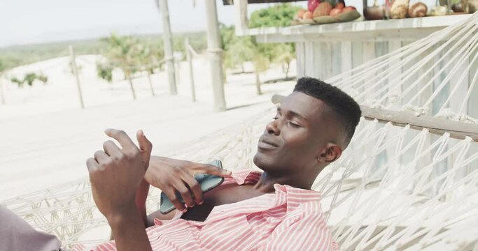 Happy african american man using smartphone lying in hammock on sunny beach, slow motion