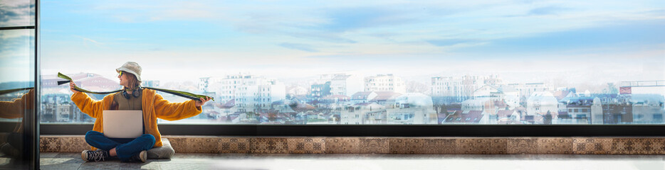 Hipster teenage girl sitting on terrace, cheerfully spreading her arms, using a laptop on her lap,...