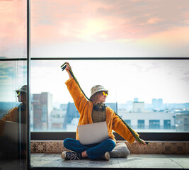 Hipster teenage girl sitting on terrace, cheerfully spreading her arms, using a laptop on her lap, with view on city and sky. Young happy woman in hat. Blank space copy and paste 