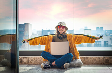 Hipster teenage girl sitting on terrace, cheerfully spreading her arms, using a laptop on her lap, with view on city and sky. Young happy woman in hat. Blank space copy and paste 