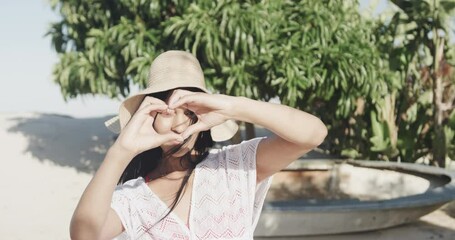 Portrait of happy hispanic woman in sunhat making heart shape with hands on sunny beach, slow motion - Powered by Adobe