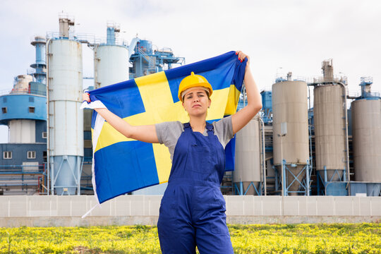 Sad female worker in hardhat with swedish flag standing in front of factory - Powered by Adobe