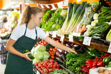 Focused fifteen-year-old girl who works part-time in a store as a trainee saleswoman puts green pepper on showcase