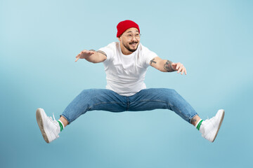 Overjoyed asian man wearing red hat, white t shirt jumping high, having fun isolated on blue background. Freedom, emotions, positive lifestyle concept
