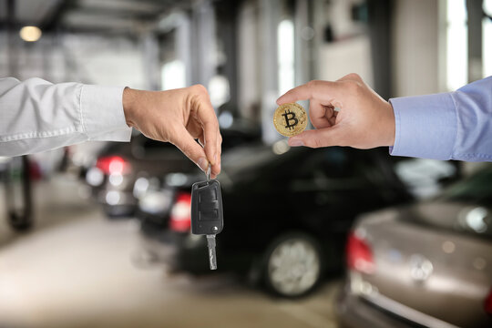Bitcoin exchange. Man using cryptocurrency to buy auto. Seller holding key and buyer with bitcoin in car dealership, closeup