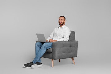 Smiling young man working with laptop in armchair on grey background