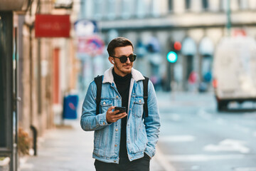A student walking through the city carrying a backpack and wearing sunglasses while using a smartphone, representing the modern urban lifestyle and reliance on technology for communication and