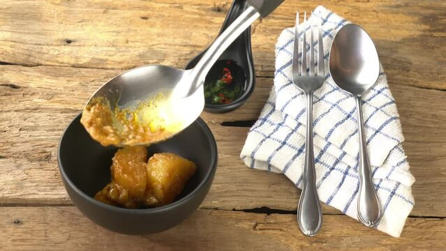 Ladling chicken curry and putting into black ceramic bowl with chilli sauce in ceramic cup and metal fork and spoon on wooden floor.