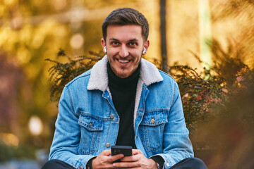 Fototapeta premium A man sitting on a bench in the city center using his smartphone and wireless headphones for a call, showcasing the convenience of technology in modern communication and the ability to multitask while