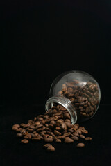 Roasted coffee beans spilling from inside a glass jar on a black background
