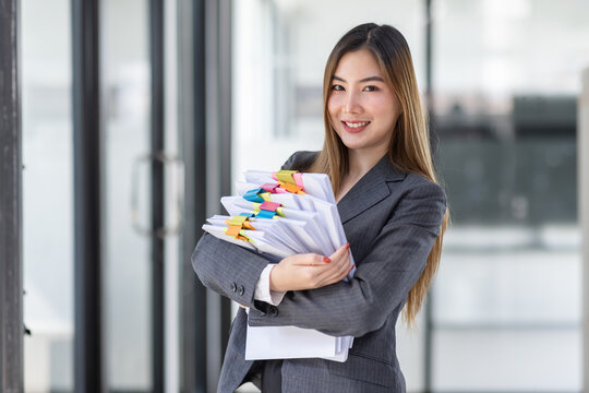 Young Business Asian Woman Holding Document File And Looking At Camera Stand In Workplace Office, Stack Of Business Overload Paper.