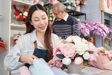Young beautiful Asian female florist entrepreneur arranging a bunch of blossoms works with old man shopkeeper, happy work in colorful flower shop store with fresh blooms, small business, family SME.