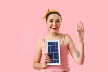 Pretty young woman with portable solar panel waving hand on pink background