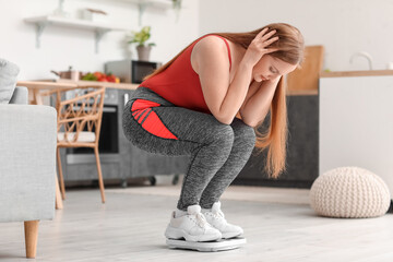 Upset young overweight woman measuring her weight on scales in kitchen