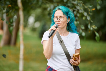 Young adult woman host of event in round glasses with turquoise dyed hair speaks into microphone at...