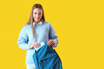 Female student putting copybook into backpack on yellow background