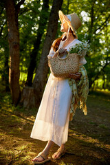 a woman in a wicker hat walks through the forest and looks towards the sunset holding a plaid and a bag in her hands