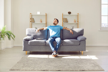Young man relaxing on couch. Man enjoys his day off or takes pause while working. Relaxed man sitting on comfortable sofa with laptop, leaning back, and enjoying good quiet peaceful time alone at home - obrazy, fototapety, plakaty