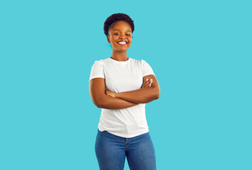 Studio shot of a happy, smiling woman in casual clothes. Portrait of a beautiful young African American girl in a white T shirt and jeans standing with her arms crossed isolated on a blue background - Powered by Adobe