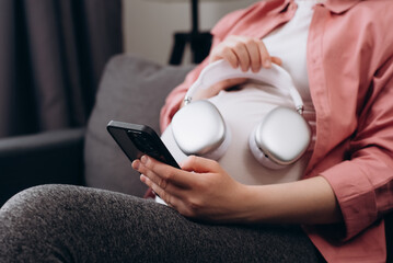 Fototapeta premium Close up of young pregnant woman applying headphones on belly sitting alone on comfy sofa at home. Expectant mother listening music in cozy living room. Future mom giving unborn listening music